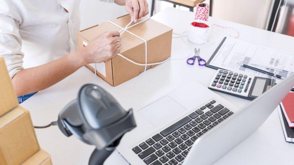 Person tying string around a cardboard box at a shipping workspace with a laptop, calculator, and barcode scanner nearby. Indicating a small business owner or employee.